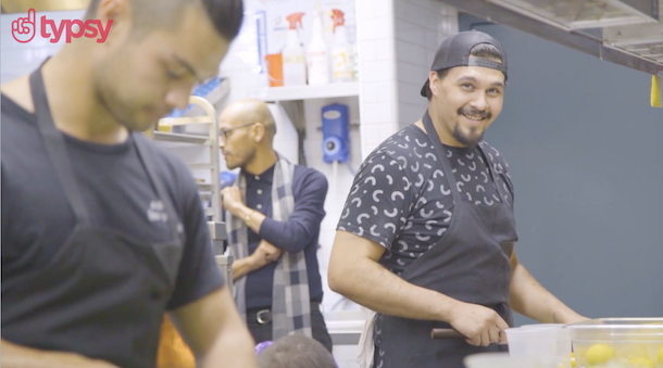 A young man with a beard and wearing a ball cap backwards works in a restaurant kitchen.