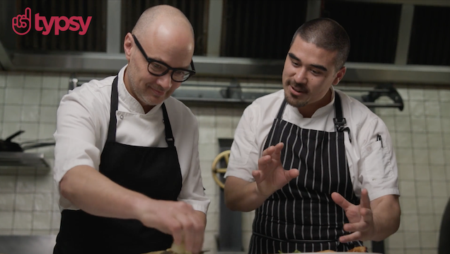 Typsy logo in red in upper left hand corner of image with two men, one wearing glasses, in chef's aprons in a commercial kitchen.