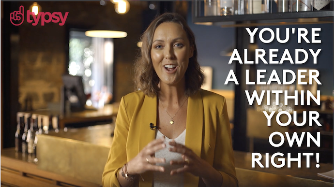 A young woman in a yellow jacket stands before a bar during a Typsy training video on leadership.