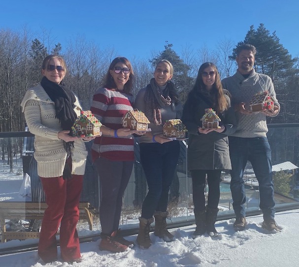 Four women and a man stand outside on a deck holding gingerbread houses with a blue sky in the background.