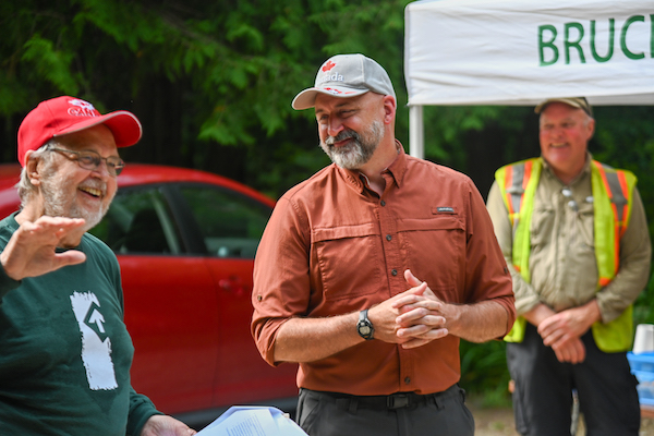 Three men stand against a background of a red car and forested area at the Celebration of Hogg's Falls on July 6, 2023.