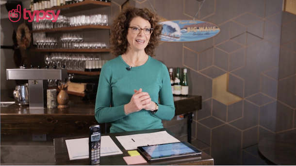 A woman in glasses and curly hair wearing a green sweater stands at a hostess desk in a restaurant.