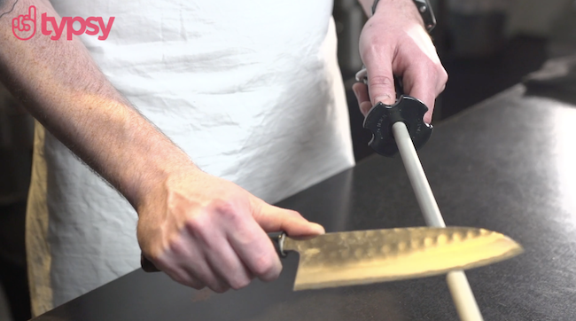 Image shows the hands of a man, who is wearing a chef's apron, sharpening a large knife.