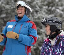 A female ski instructor wearing a blue and red ski jacket shares a laugh with a young student wearing a multi-coloured coat.